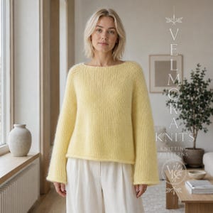 Op de afbeelding: Een lichtgele trui met lange mouwen, een ronde halslijn en een losse, ontspannen pasvorm. De gebreide stof heeft een zachte, pluizige textuur. De tekst VELMORA KNITS KNITTING PATTERNS is zichtbaar aan de rechterkant van de afbeelding.