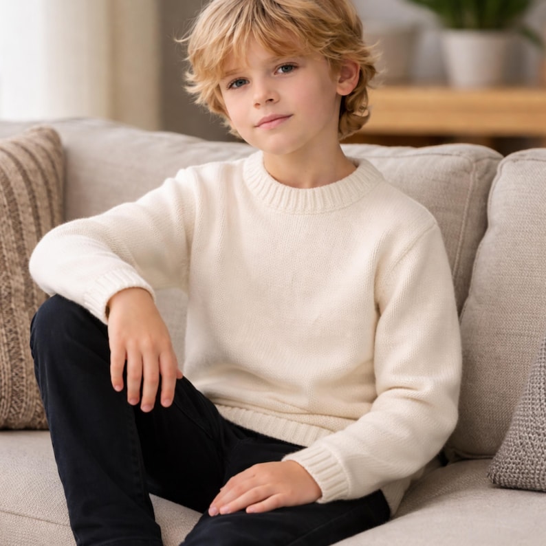 May include: A cream-colored sweater with a crew neck. The sweater is worn by a person sitting on a beige sofa. The person is also wearing dark blue pants. The background includes a patterned pillow and a wooden table.