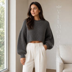 Könnte beinhalten: Ein anthrazitfarbener, bauchfreier Pullover mit gerippter Strickstruktur, langen Ballonärmeln und Rundhalsausschnitt. Der Text VELMORA KNITS KNITTING PATTERN ist rechts im Bild in einer dünnen, weißen, vertikalen Serifenschrift zu sehen.
