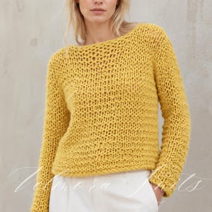 Könnte beinhalten: Ein senfgelber Langarmpullover mit einer lockeren, grob gestrickten Struktur und einem weiten U-Boot-Ausschnitt. Das Kleidungsstück wird über einer schlichten weißen Hose getragen. Das Strickmuster ist über den gesamten Körper und die Ärmel einheitlich.