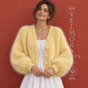May include: A pale yellow fuzzy knit cardigan with long voluminous sleeves, a V-neckline, and a front button closure. The garment is worn over a white dress. Text on the right side reads VELMORA KNITS.