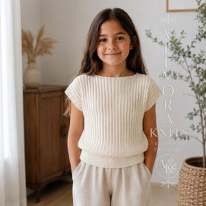 Pode incluir: Uma blusa de malha cor de creme de manga curta com textura canelada vertical, decote canoa largo e bainha ajustada. A peça é exibida com o texto sobreposto VELMORA KNITS KNITTING PATTERNS e um ícone de logótipo.