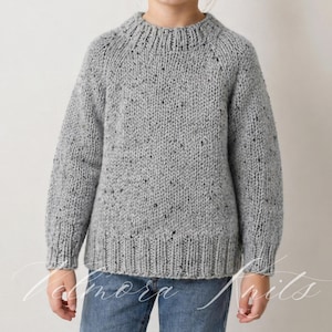 Puede incluir: Un suéter de punto gris con un patrón moteado. El suéter tiene cuello, puños y dobladillo de canalé. Las mangas son largas y el suéter se usa con vaqueros azules. El texto "Tednora Knits" es visible en la esquina inferior izquierda.