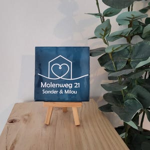 Op de afbeelding: Een donkerblauwe vierkante tegel met witte tekst en een huis- en hartafbeelding, gepresenteerd op een klein houten ezel. De tekst luidt "Molenweg 21 Sander & Milou."