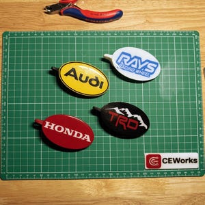 May include: Oval car emblem keyrings featuring the logos of Audi, Honda, TRD, and Rays Racing Wheel. The keyrings are in yellow, red, black, and white. A green cutting mat and tweezers are also visible.