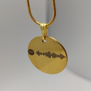 Könnte beinhalten: Eine goldfarbene Halskette mit Anhänger und einem kreisförmigen Anhänger. Der Anhänger zeigt ein schwarzes Spotify-Code-Design. Die Halskette ist ebenfalls goldfarben und hängt vor einem weißen Hintergrund.