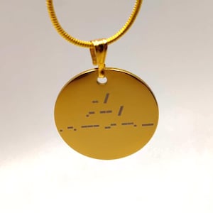 Op de afbeelding: Een goudkleurige ketting met een cirkelvormige hanger. De hanger heeft gegraveerde morsecode symbolen. De ketting is een dunne, goudkleurige slangenketting. De ketting wordt getoond tegen een witte achtergrond.