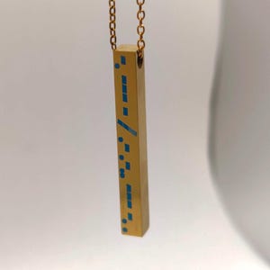 May include: A gold-colored pendant necklace with a rectangular bar pendant. The bar features a pattern of blue squares and lines. The necklace chain is also gold-colored. The pendant hangs vertically.