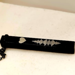 May include: A black rectangular pendant necklace with a silver heart and sound wave design. The pendant is suspended from a black chain. The necklace is a piece of jewellery.