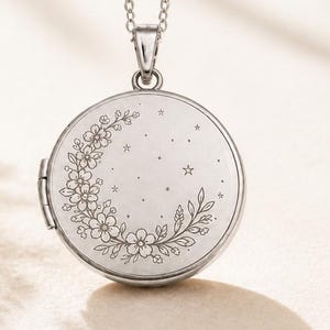May include: A silver-toned circular locket pendant suspended from a delicate matching chain. The front surface of the locket features a fine-line engraved design of a crescent moon formed by floral blossoms and leaves, accented with small stars.