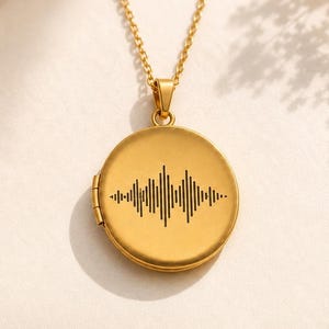 May include: A gold-toned circular locket pendant suspended from a matching delicate chain. The front surface of the locket features a black engraved sound wave pattern centered horizontally. The metal has a smooth, matte finish throughout.