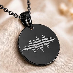 Pode incluir: Um colar com pendente circular preto apresentando um padrão de onda sonora gravado em branco na parte frontal. O pendente está preso a uma corrente de metal preto a condizer, criando um visual elegante e monocromático com um gráfico de áudio.