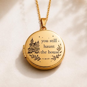 Può includere: Un ciondolo a medaglione circolare color oro su una catenina abbinata. La parte anteriore del medaglione presenta un'illustrazione incisa di una casa e il testo, you still haunt the house, circondato da piccoli dettagli a foglia e stelle.