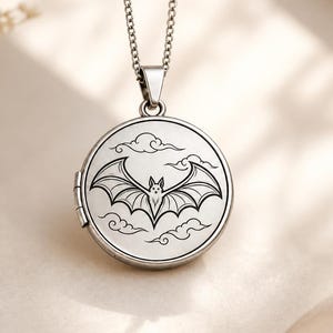 May include: A round silver-toned metal locket pendant suspended from a matching link chain. The front of the locket features a black engraved illustration of a bat with its wings spread wide, surrounded by stylized cloud motifs against a smooth surface.
