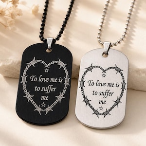 Puede incluir: Dos collares con placas de identificación, uno negro mate y otro plateado, cada uno con un corazón grabado con láser hecho de alambre de púas y el texto "To love me is to suffer me" en el centro. Ambos colgantes cuelgan de cadenas de bolas.