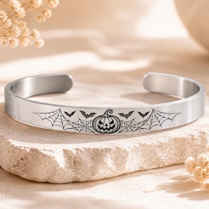Könnte beinhalten: Ein silberfarbenes Metall-Armband mit einer schwarzen Gravur, die einen zentralen Halloween-Kürbis zeigt, umgeben von detaillierten Spinnennetzen und kleinen fliegenden Fledermäusen, mit einer offenen Schiene zur Anpassung.