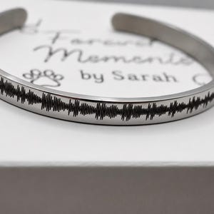 Peut inclure: Un bracelet manchette argenté avec un motif d'onde sonore noir gravé sur le pourtour. Le bracelet est posé sur une surface blanche avec le texte "Forever Moments by Sarah".