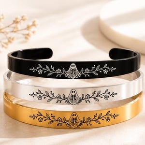 May include: Three cuff bracelets stacked vertically, featuring a black, a silver-tone, and a gold-tone finish. Each bracelet is engraved with a central ghost figure adorned with a bow, flanked by symmetrical floral and leafy vine patterns.