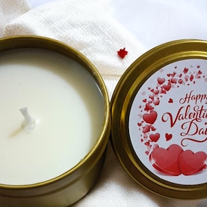 Puede incluir: Una lata de vela dorada con una vela blanca y una mecha. La tapa, colocada a un lado, presenta un diseño con corazones rojos y el texto "Happy Valentine's Day". La vela está sobre un paño blanco.