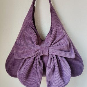 Puede incluir: Bolso de hombro de pana morado con un gran lazo. El bolso tiene una forma estructurada con una correa ancha y un lazo prominente en la parte delantera. El bolso está hecho de una tela suave y acanalada.