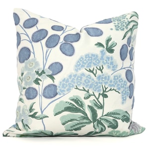 May include: Decorative throw pillow with a floral pattern. The pillow features a white background with blue and green botanical illustrations. The design includes large blue oval shapes, clusters of blue flowers, and green leaves. The pillow is square and appears to be made of fabric.