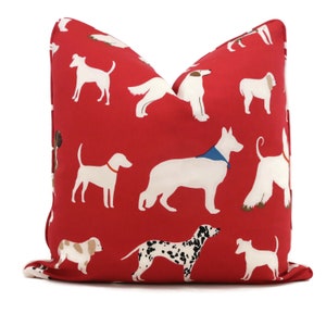 以下が含まれることがあります： 赤色の装飾用クッション。白い犬のイラストが繰り返されるパターンです。犬は様々なポーズで描かれており、中には色のついた首輪やバンダナをしているものもあります。クッションは正方形です。