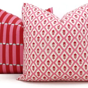 May include: Two decorative throw pillows with pink and red geometric patterns. The pillow on the left has a striped pattern with a repeating design of circles. The pillow on the right has a diamond pattern with a repeating design of stylised flowers.