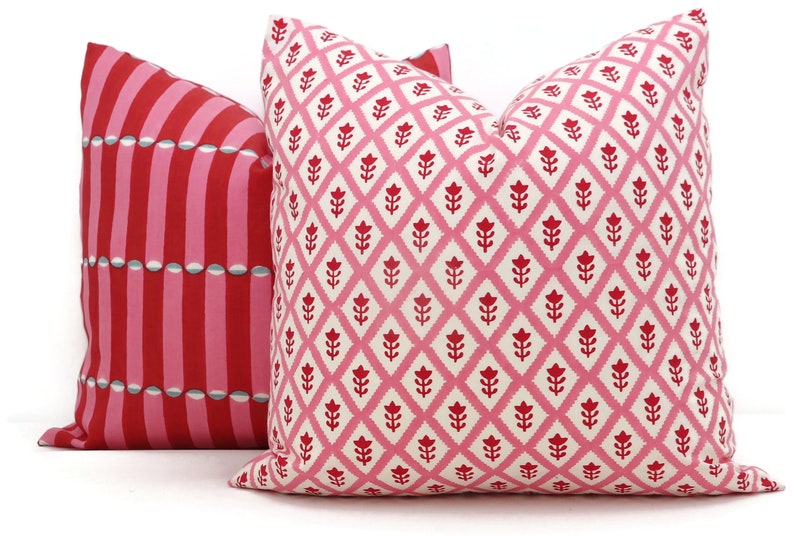 May include: Two decorative throw pillows with pink and red geometric patterns. The pillow on the left has a striped pattern with a repeating design of circles. The pillow on the right has a diamond pattern with a repeating design of stylised flowers.