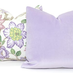 May include: Two decorative throw pillows, one with a floral pattern in shades of pink, green, and purple on a white background, and the other a solid lavender colour.