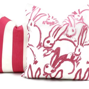 Puede incluir: Dos almohadas decorativas. Una almohada es blanca con un patr&oacute;n de conejo rosa. La otra almohada es blanca con rayas rosas.