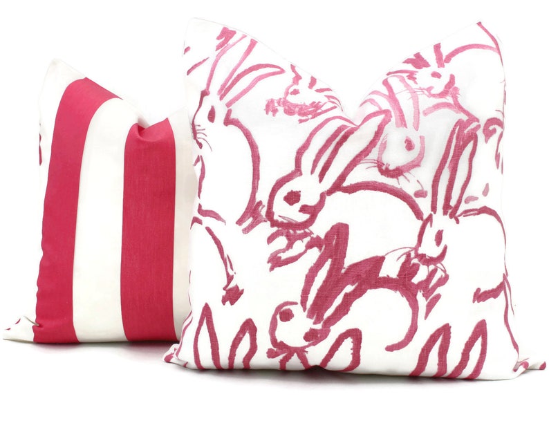 Puede incluir: Dos almohadas decorativas. Una almohada es blanca con un patr&oacute;n de conejo rosa. La otra almohada es blanca con rayas rosas.