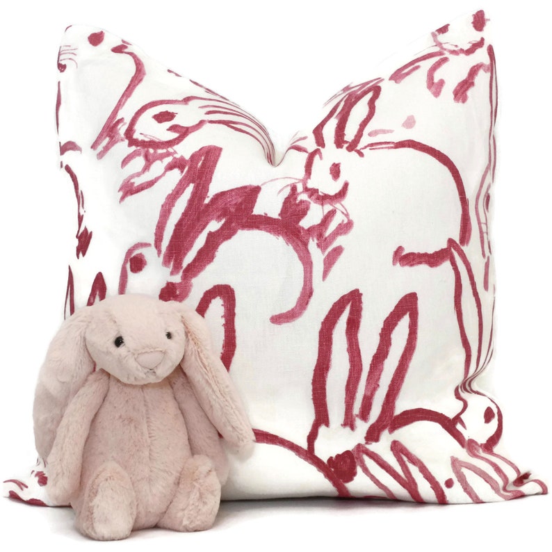 Puede incluir: Una almohada blanca con un dise&ntilde;o de conejo rosa. Un conejo de peluche rosa est&aacute; sentado frente a la almohada.