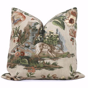 May include: Decorative throw pillow with a pastoral scene featuring a deer, a stone structure, and floral elements in shades of brown, green, and red on a beige background. The pillow has a soft, textured appearance.