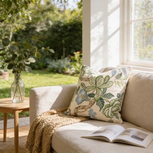 a cozy living room with a beige sofa, a wooden side table, and a vase of greenery. A large pillow with a floral pattern rests on the sofa, and an open book lies on the armrest. The room is bathed in natural light from a large window, and the garden outside is visible, creating a serene and inviting atmosphere.