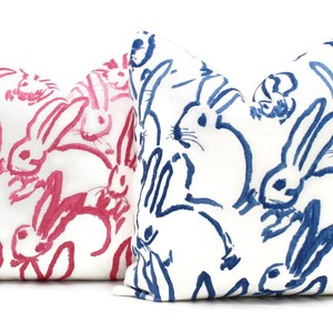 Puede incluir: Dos almohadas decorativas con fondo blanco y un patr&oacute;n de conejos rosas y azules. Los conejos est&aacute;n dibujados de forma simple y estilizada.