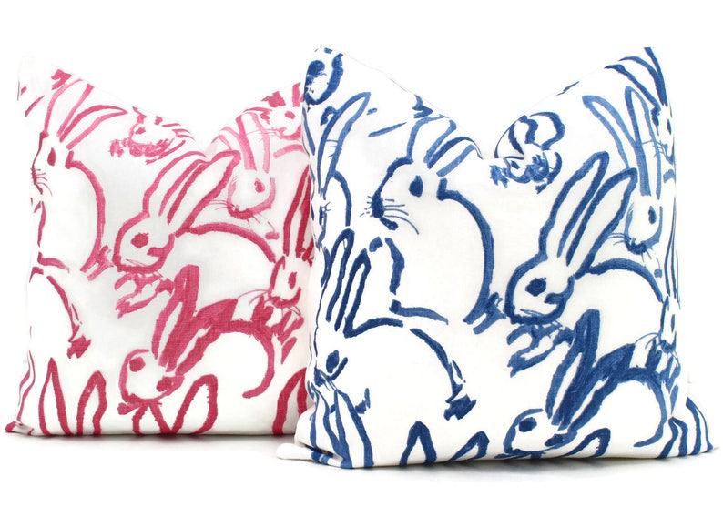 Puede incluir: Dos almohadas decorativas con fondo blanco y un patr&oacute;n de conejos rosas y azules. Los conejos est&aacute;n dibujados de forma simple y estilizada.