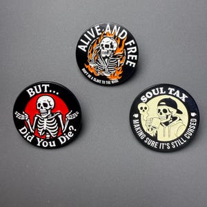 Könnte beinhalten: Drei schwarze runde Buttons mit Skelettgrafiken. Einer sagt "ALIVE AND FREE" mit einem Skelett in Flammen. Ein anderer sagt "BUT... Did You Die?" mit einem Skelett. Der dritte sagt "SOUL TAX" mit einem Skelett.