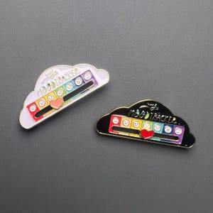 May include: Two enamel pins shaped like clouds, each with a mood tracker design. One has a white cloud outline, the other is black. Both feature a rainbow slider, a red heart, and the text "MOOD TRACKER".