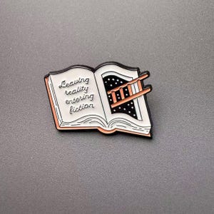 May include: A black and white enamel pin shaped like an open book. The left page features the text 'Leaving reality entering fiction' in black script. The right page shows a black starry night sky with a small ladder extending from the book pages.