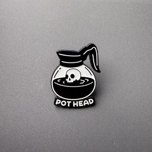 May include: Black and white enamel pin featuring a coffee pot design with a skull inside. The pin has the words "POT HEAD" in white text at the bottom. The pin is on a gray background.