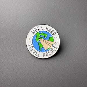 May include: Round enamel pin with a white border, featuring a blue and green globe with a paper airplane. The pin has the text "WORK HARD TRAVEL HARDER" in black lettering.