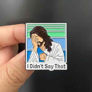 May include: A rectangular enamel pin featuring an illustration of a figure with long brown hair and a beard, covering their face with their hand. The pin has a blue, green, and white background and the text "I Didn't Say That".