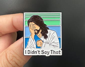 May include: A rectangular enamel pin featuring an illustration of a figure with long brown hair and a beard, covering their face with their hand. The pin has a blue, green, and white background and the text "I Didn't Say That".