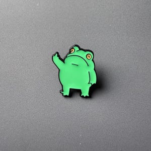 Puede incluir: Un pin de esmalte con forma de rana verde con un contorno negro. La rana está saludando con un brazo levantado y tiene ojos amarillos. El pin está sobre un fondo gris.