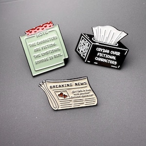 Peut inclure: Trois broches en émail sur le thème de la culture populaire. L'une est une note vert clair à pois rouges avec le texte : NOTE THE CHARACTERS ARE FICTION. THE EMOTIONAL DAMAGE IS REAL. Une autre est une boîte à mouchoirs noire avec le texte : CRYING OVER FICTIONAL CHARACTERS. La troisième est un journal avec le texte : BREAKING NEWS Girl falls in love with another fictional character.