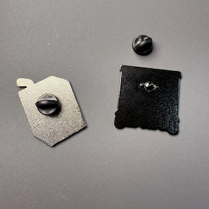 May include: Two enamel pins with black backs and pin clasps. One pin is a silver-colored tag shape, and the other is a black square shape. A separate black pin clasp is also visible. The pins are on a gray surface.