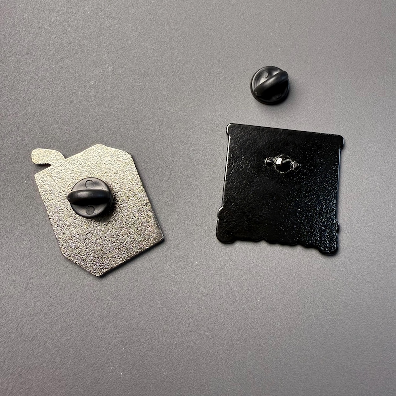 May include: Two enamel pins with black backs and pin clasps. One pin is a silver-colored tag shape, and the other is a black square shape. A separate black pin clasp is also visible. The pins are on a gray surface.