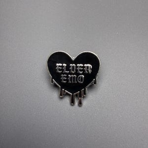 Op de afbeelding: Een zwarte hartvormige emaille speld met zilverkleurige metalen randen. De tekst ELDER EMO staat in een zilverkleurig gotisch lettertype in het midden. De onderkant van het hart heeft een druipend vloeistofeffect met zilverkleurige metalen omlijningen.