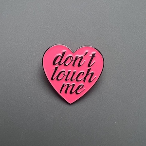 Puede incluir: Un pin de esmalte en forma de corazón rosa con las palabras "don't touch me" en letras cursivas negras. El pin tiene un borde negro y está sobre un fondo gris. Un accesorio de declaración.