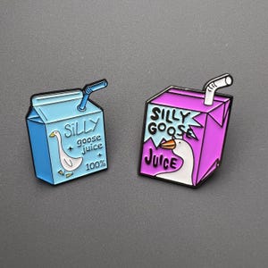 Pode incluir: Dois pinos de esmalte em forma de caixas de sumo. Um é azul claro com uma ilustração de ganso e as palavras "SILLY goose juice 100%". O outro é roxo com um ganso e as palavras "SILLY GOOSE Juice". Ambos têm uma palhinha branca.